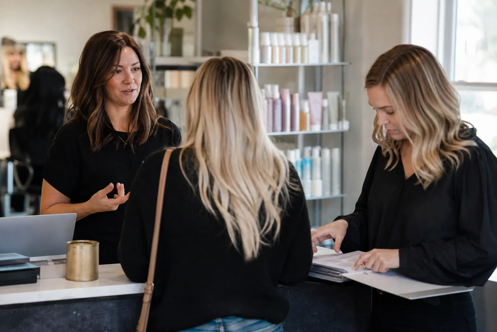 A salon reception area with a stylist speaking with a client while another staff member checks a paper schedule at the counter, with product shelves and soft daylight in the background.