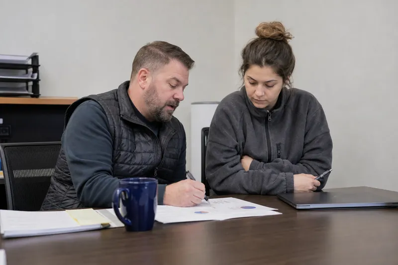 A service business owner and a team member sit at a small desk in a plain office, looking over a printed monthly report with pens, coffee, and a laptop closed beside them.