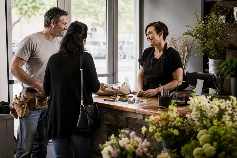 A local contractor and a salon owner chatting with a customer at a small storefront counter, with tools and fresh flowers nearby, in natural daylight.