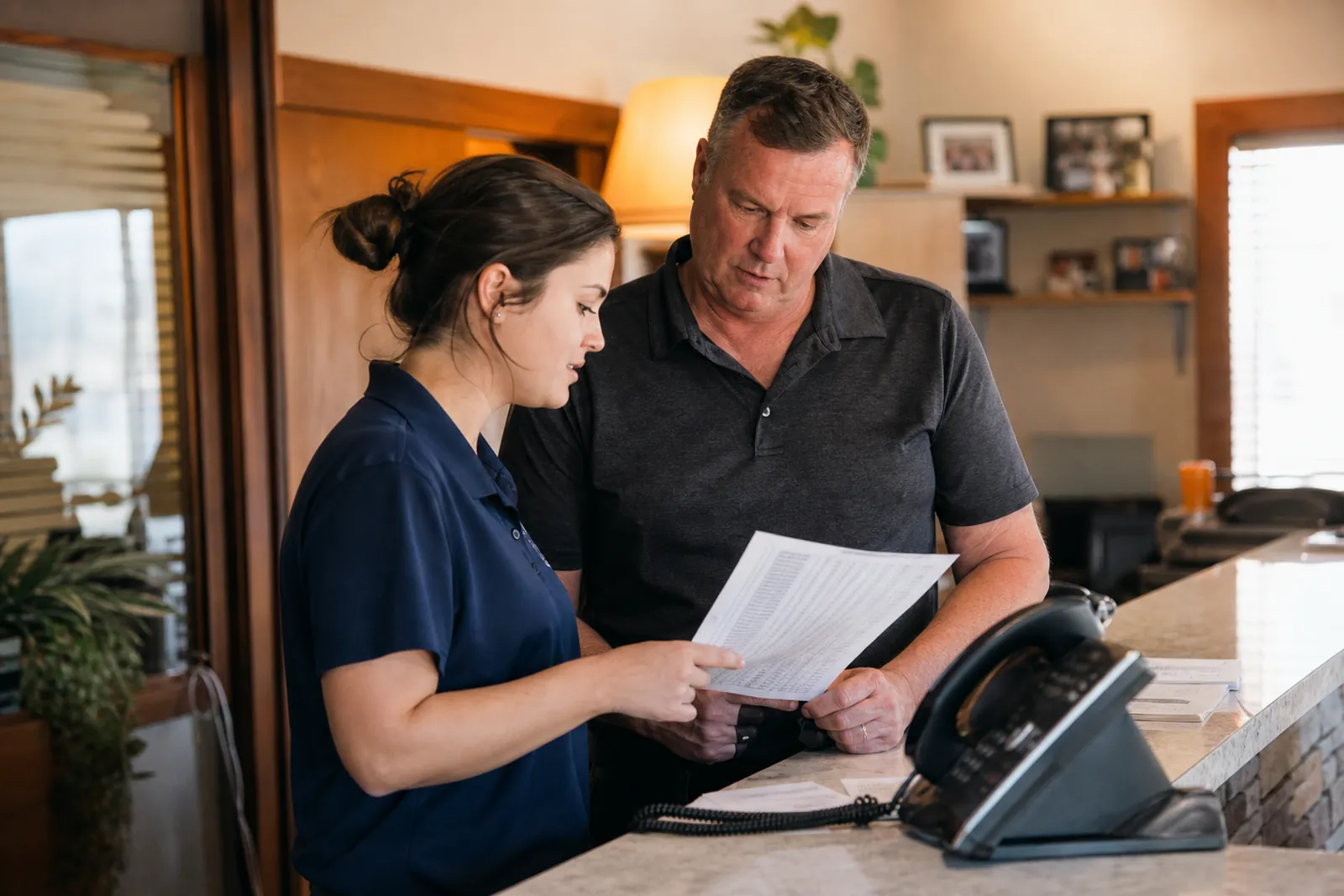 A service business owner and a front-desk staff member standing near the counter in a small office, looking at a printed call log while a phone sits nearby, with warm natural light and a lived-in, working atmosphere.