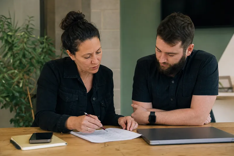 A small business owner and a web studio teammate sit at a table in a simple office, looking over a printed checklist with a laptop closed beside a phone and a notebook.