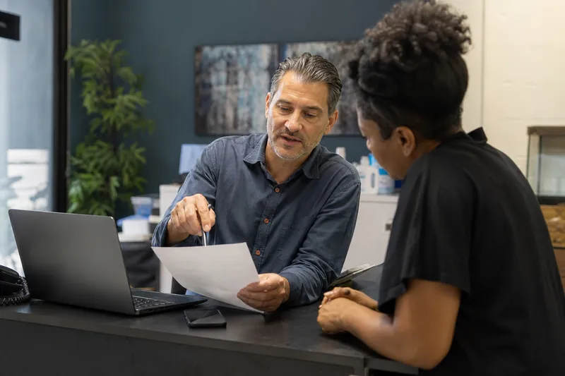 A small business owner in a simple office with a laptop and phone beside them, reviewing a printed contact form while talking with a staff member near a front desk.