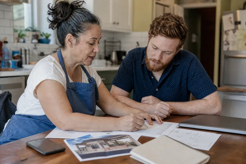 A small business owner and a web helper sitting at a kitchen table with a notebook, a laptop closed to the side, and printed account records spread out, while a storefront photo an