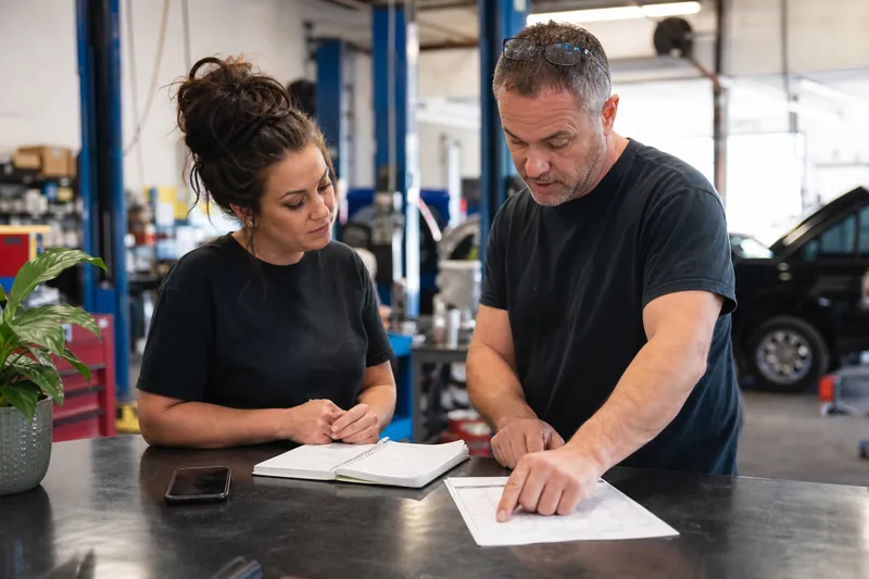 A small business owner and a team member standing by a front counter with a printed calendar, a notebook, and a phone, reviewing renewal dates in a busy service shop.