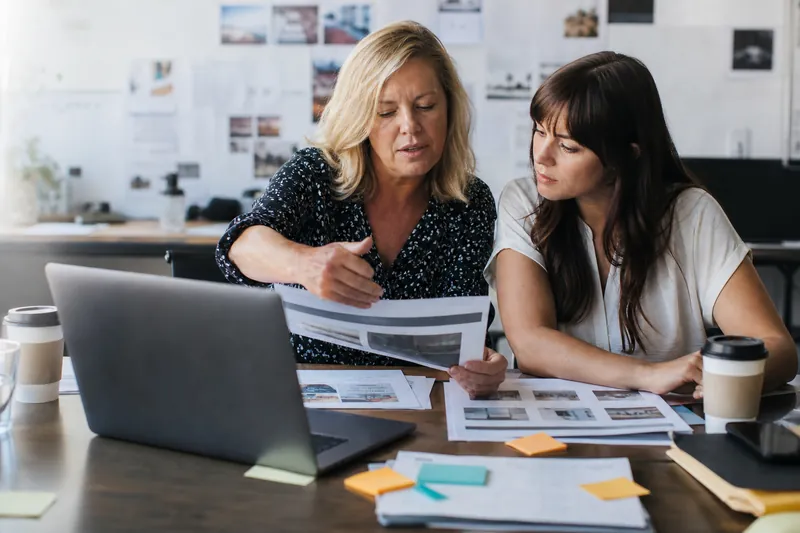 A small business owner and a web helper sit at a cluttered desk in a bright office, reviewing printed website pages beside a laptop, with coffee cups and sticky notes spread around