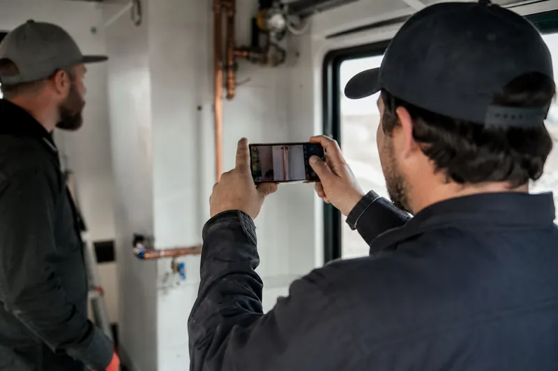 A service worker in a neat work truck or job site room holds a phone with both hands while filming a finished repair, with another person nearby and natural side light coming throu