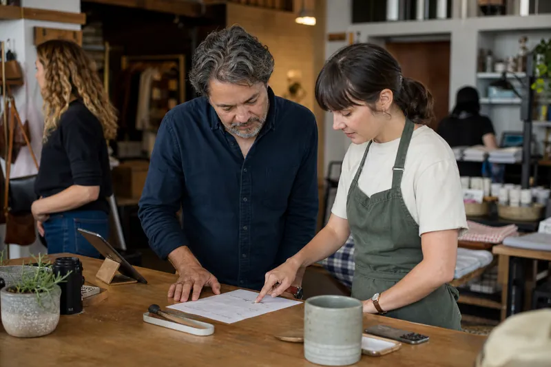A small business owner and a team member standing at a workbench in a tidy local shop, looking over a printed monthly notes sheet while customers and tools sit naturally in the bac