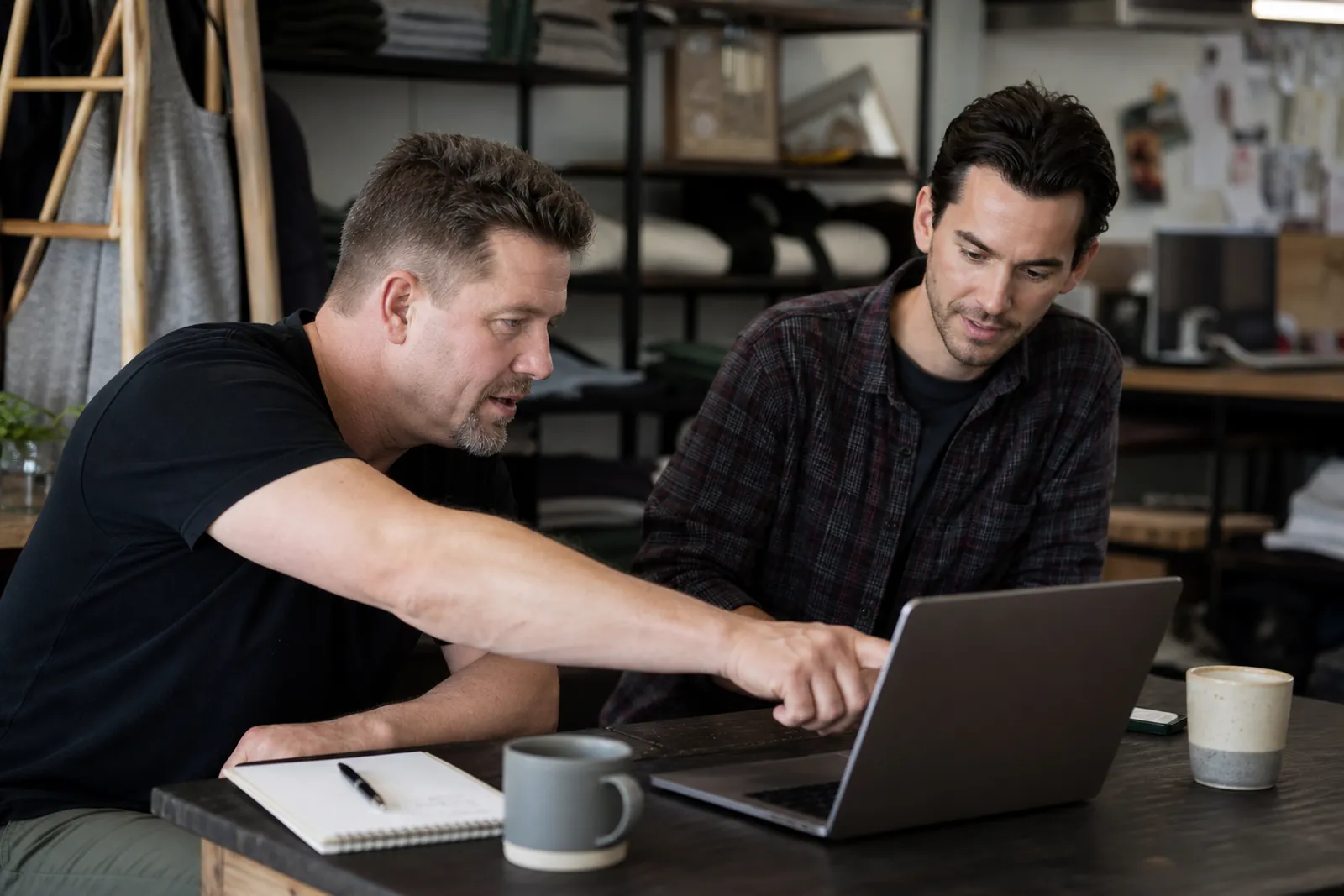 A business owner and a web helper sitting at a small desk in a shop office, looking over a laptop together with a notepad and coffee cups nearby, in a candid natural moment.