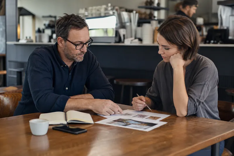A small business owner and a web designer sit across a table in a coffee shop with a printed proposal, a notebook, a phone, and a few business photos spread out between them.
