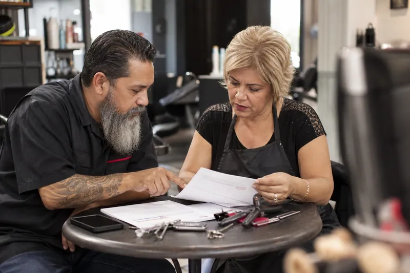 A locksmith and a salon owner sit together at a small table in a quiet neighborhood shop, reviewing printed search reports while tools, styling combs, and everyday work items sit n