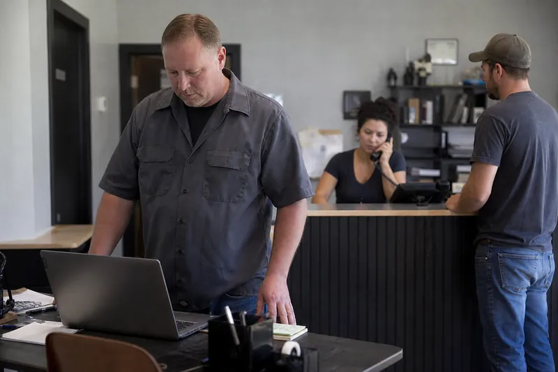 A small business owner in work clothes stands beside a desk with a laptop open, while a teammate answers a phone in the background and a customer waits near the counter in a modest