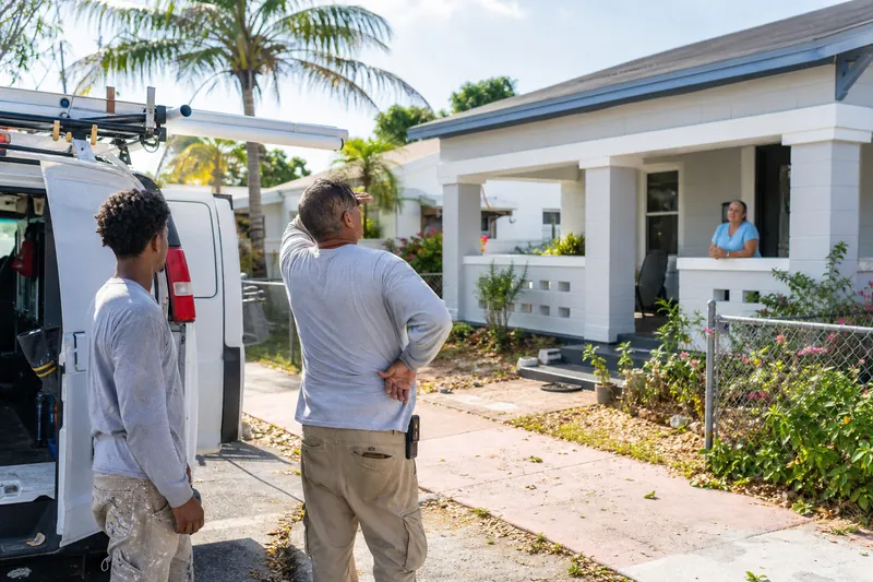 A candid photo of a small service crew standing on a quiet residential street after finishing work, with one person holding tools and another talking to a homeowner near a truck in
