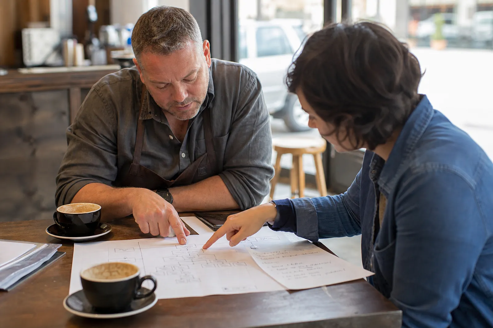 A business owner and a web designer sit at a small table in a local shop, pointing at a printed site map and handwritten service notes while coffee cups sit nearby.