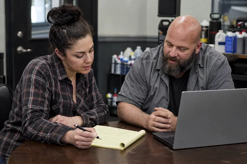 A local business owner and a web helper sit at a small table in a shop office, reviewing handwritten page titles on a notepad beside a laptop, with tools and service supplies in th