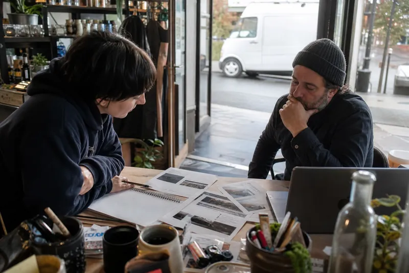 A small team member and a business owner sit at a cluttered desk in a local shop, looking over printed website pages and a notebook while the open front door shows a parked service