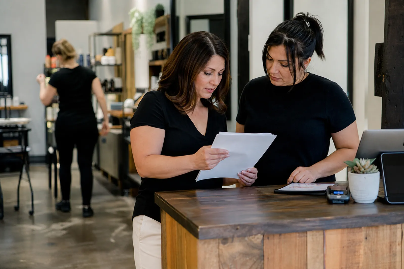 A small service-business owner and a staff member stand near the front counter of a tidy local shop, looking over printed notes together while the work area sits active in the background.