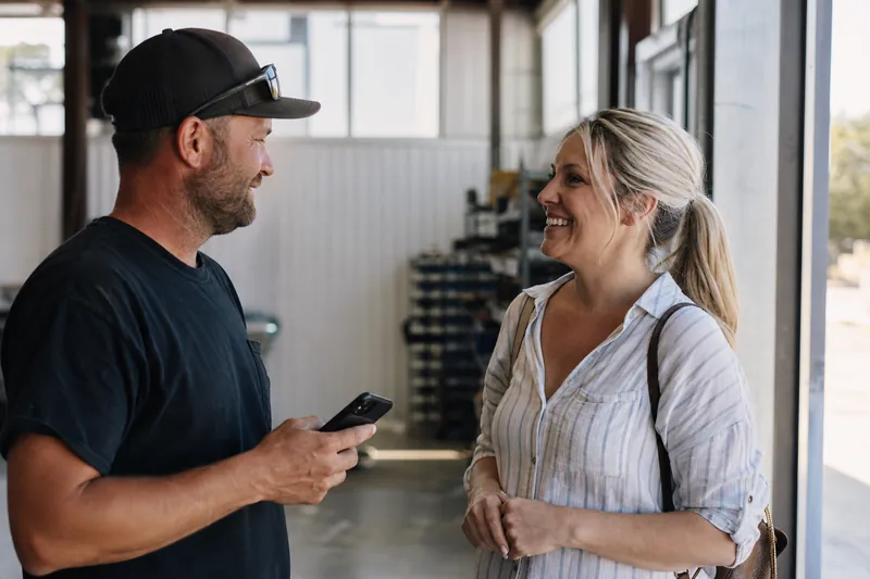 A small service business owner and a smiling customer standing near the finished work in a bright shop or job site, with a phone held at chest height during a casual conversation.