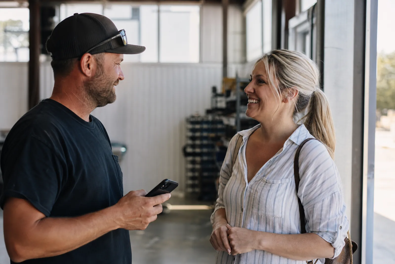 A small service business owner and a smiling customer standing near the finished work in a bright shop or job site, with a phone held at chest height during a casual conversation.