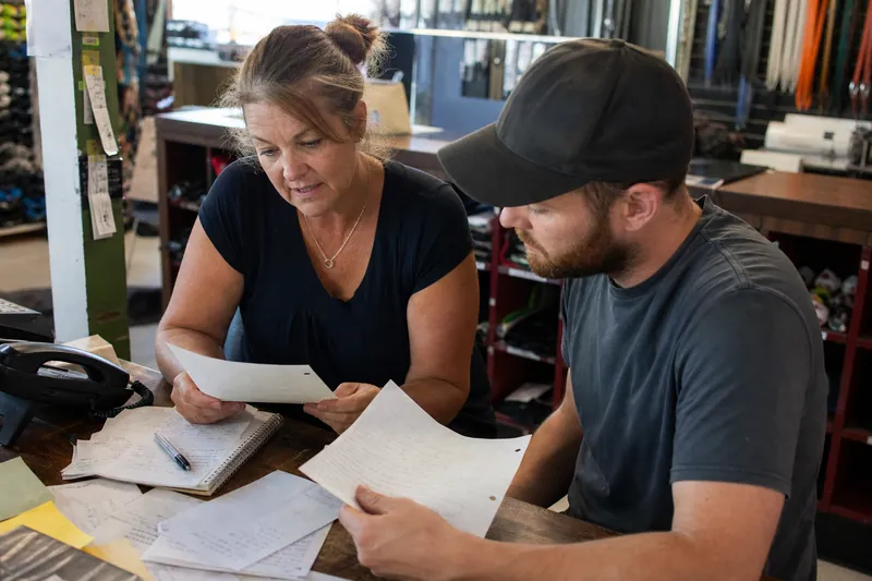 A local business owner and a team member sit at a small desk in a shop or office, sorting handwritten customer questions on paper beside a phone and a notebook, with a working coun