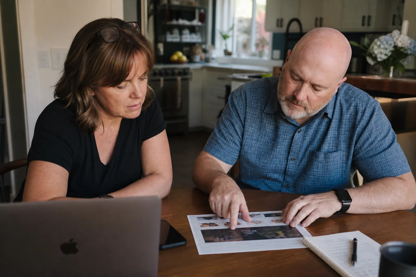 A small business owner sits at a kitchen table with a laptop and a phone beside a notepad, while a second person points at a printed website draft during a casual review.
