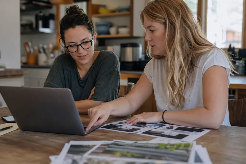 A small business owner and a web designer standing in a modest office with a laptop open, reviewing a website plan over a notebook and phone, with natural light and a casual workin