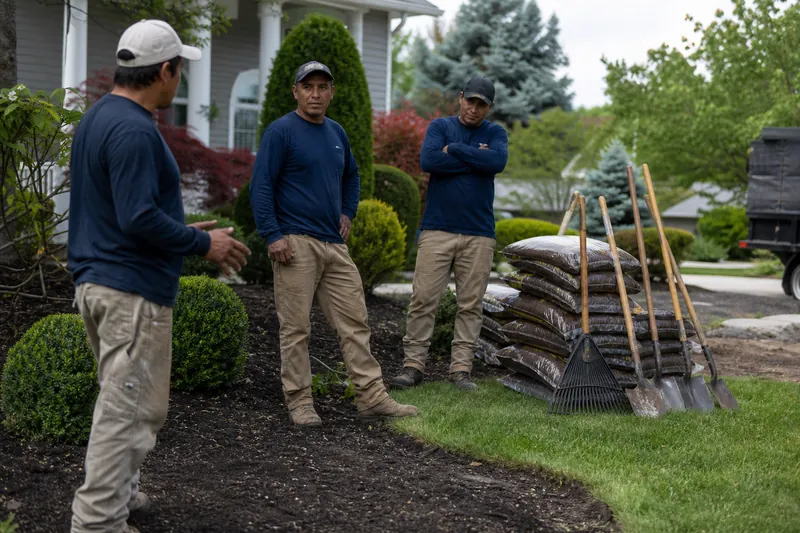 A landscaper in a work truck is talking with a homeowner beside a neatly trimmed front yard, with tools and fresh cut grass visible in the driveway.