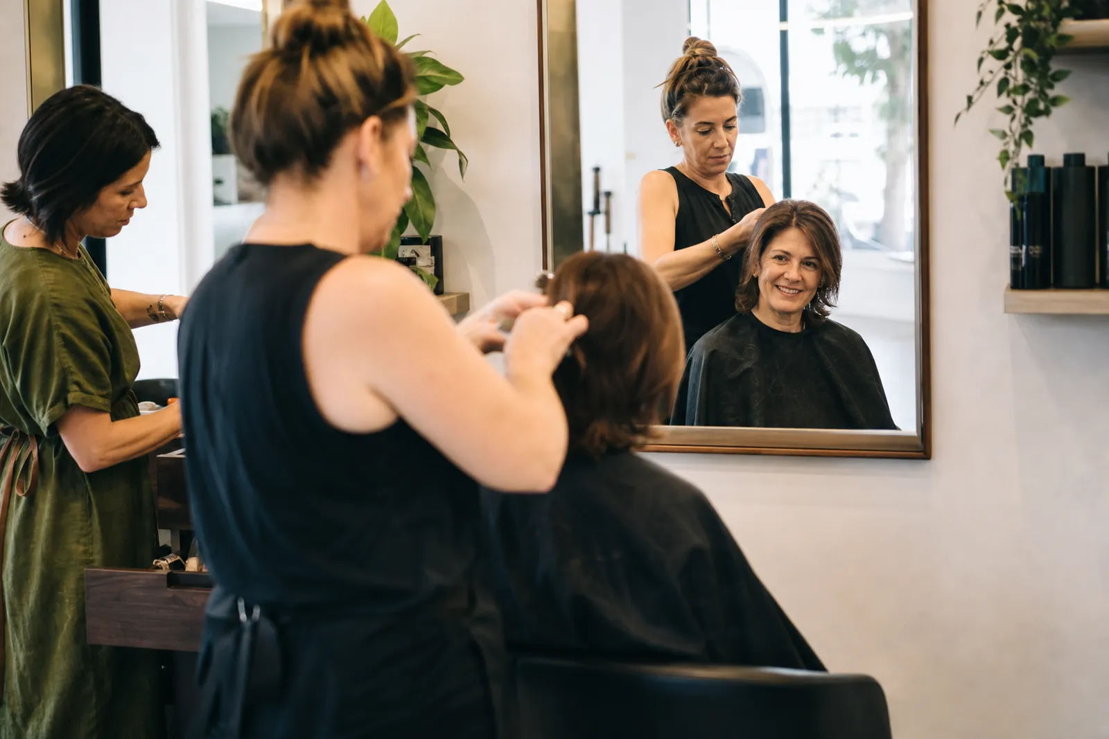 A candid photo inside a bright neighborhood salon with a stylist finishing a haircut in the chair while another team member tidies tools nearby and a client smiles in the mirror.