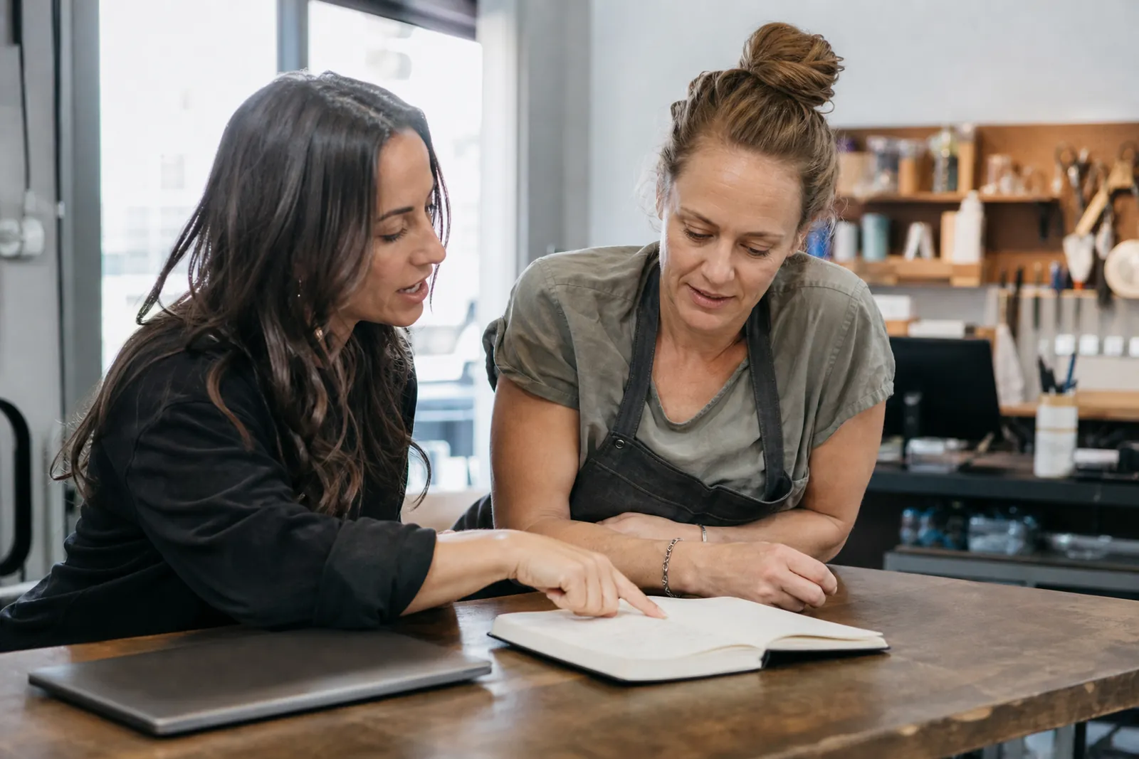A small business owner and a web helper sitting at a counter in a quiet shop, reviewing a notebook with a laptop closed nearby while a service desk and everyday tools sit in the background.