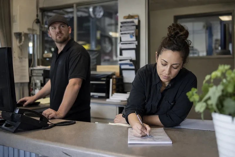 A small office counter in a service business with two staff members near a ringing phone, one person glancing up while another finishes a note on paper, with tools and work orders 