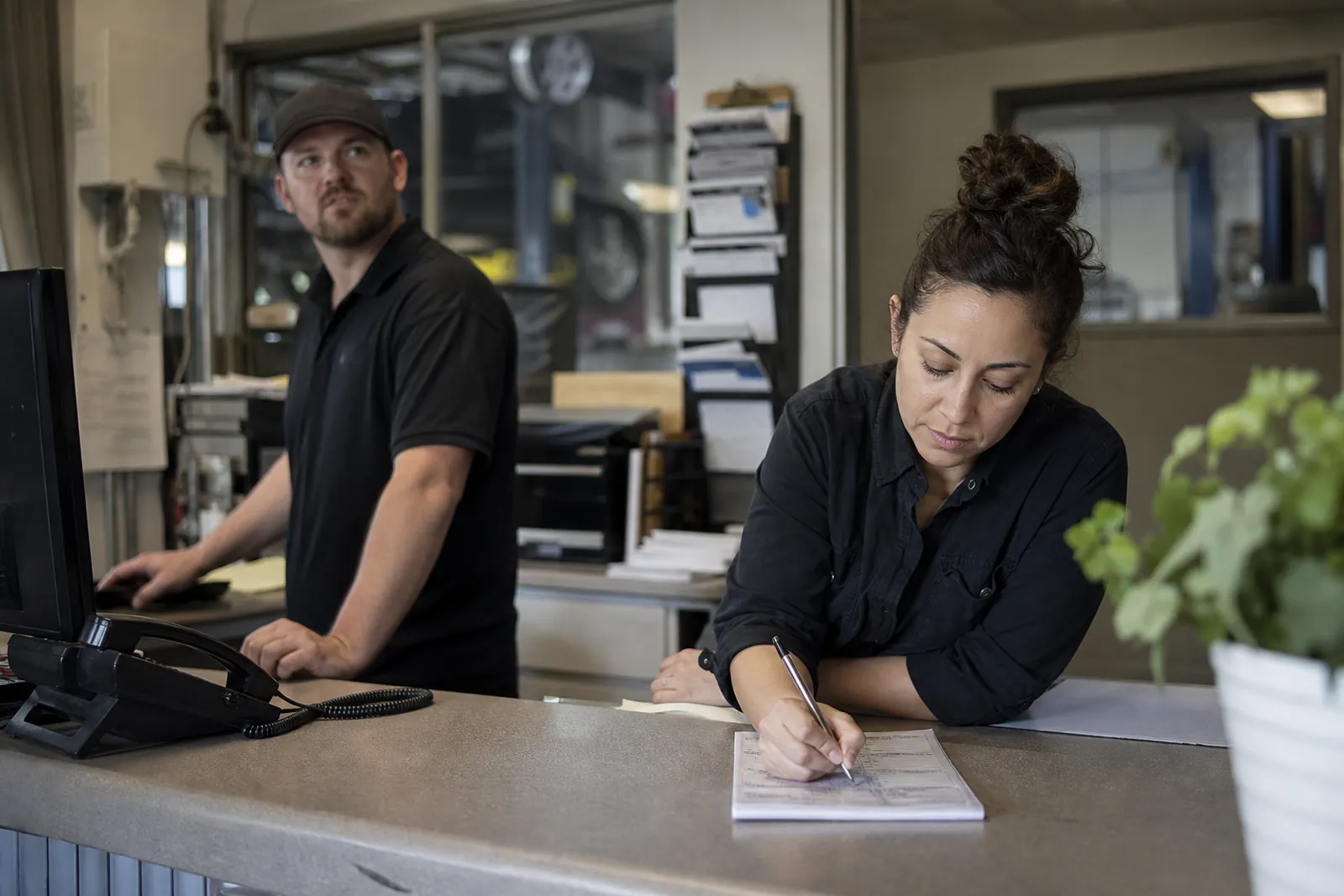 A small office counter in a service business with two staff members near a ringing phone, one person glancing up while another finishes a note on paper, with tools and work orders in the background.