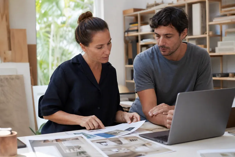 A small business owner and a web designer stand in a bright Miami studio reviewing printed photos and a laptop mockup on a desk, with soft afternoon light and shelves of project ma