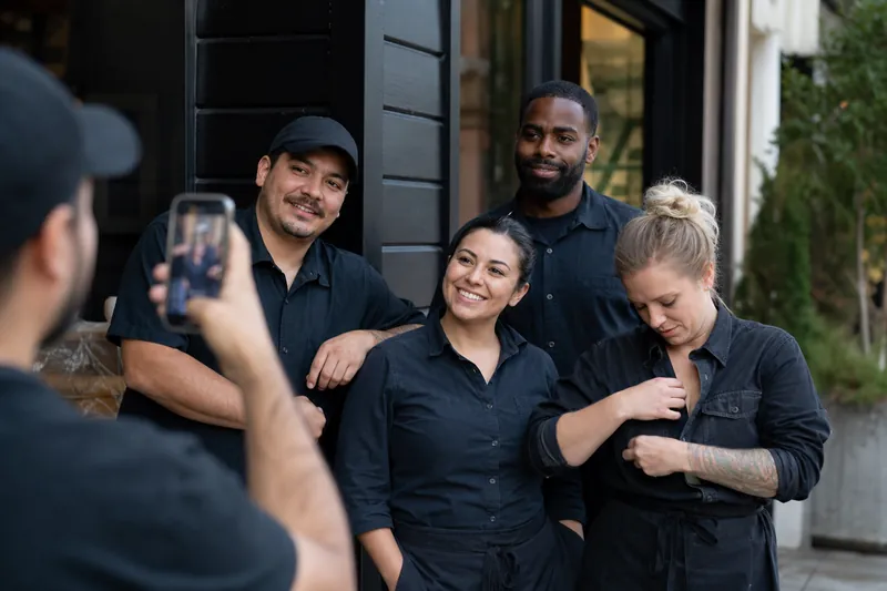 A small service crew stands outside a neighborhood business during a quiet morning, with one person adjusting a work shirt while another holds a phone for a casual team photo.