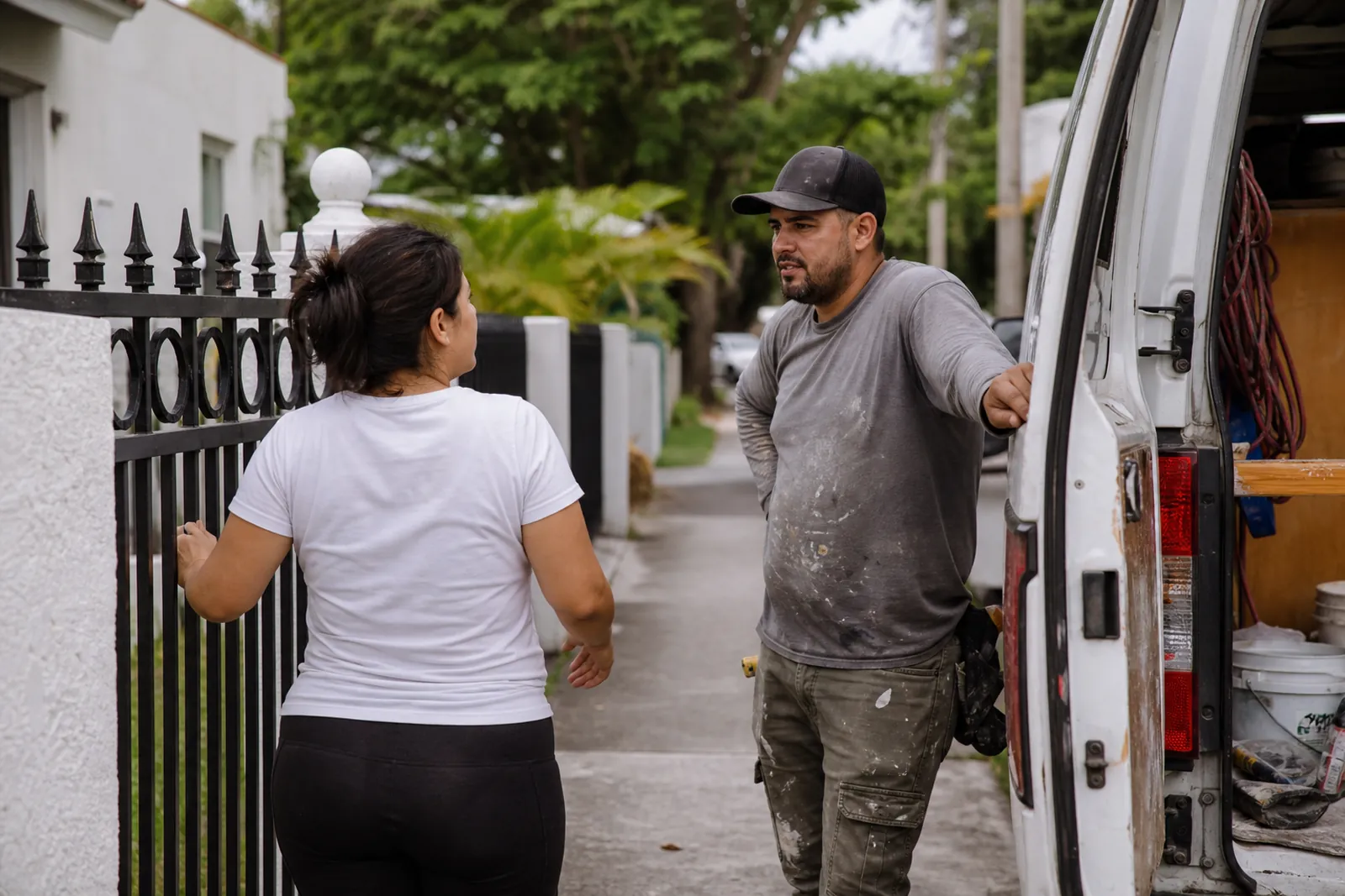 A contractor and a homeowner standing together at the front gate of a small house on a quiet Miami street, talking beside a work van with tools visible in the open back.