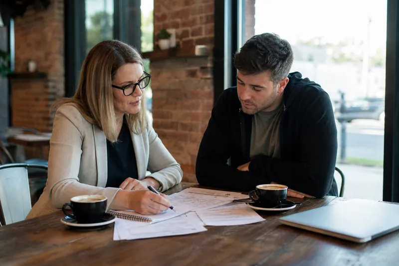 A small business owner and a web helper sitting at a cafe table with a notebook, a laptop closed beside them, and printed account notes spread out between coffee cups.