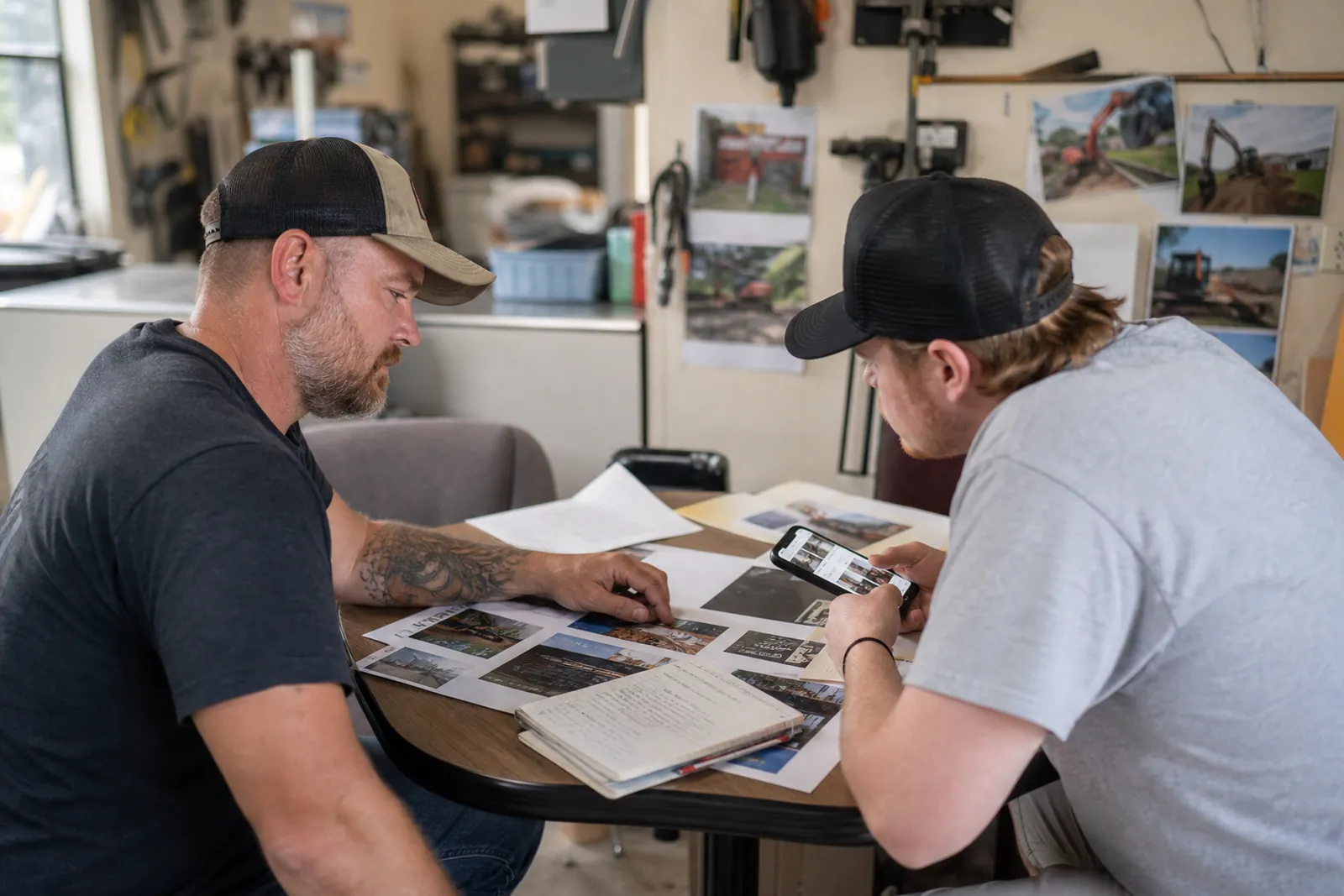 A local service owner and a teammate sitting at a small table in a shop office, looking over printed website pages, a notebook, and a phone with a business profile open, with tools and job photos visible in the backgroun