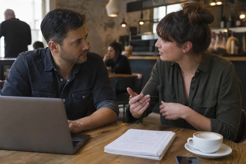 A small business owner and a web professional sit at a café table with a laptop open beside a notebook full of page notes, talking through a website plan.