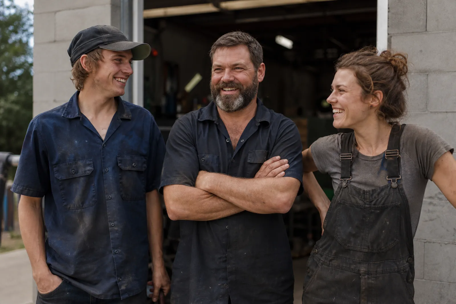 A candid photo of a small service business owner and two team members standing outside their shop entrance in daylight, wearing work clothes and smiling naturally.