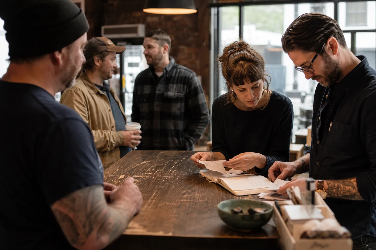 A service owner and a team member at a shop counter sorting paper customer cards and a notebook while a few regular customers wait nearby and chat casually.