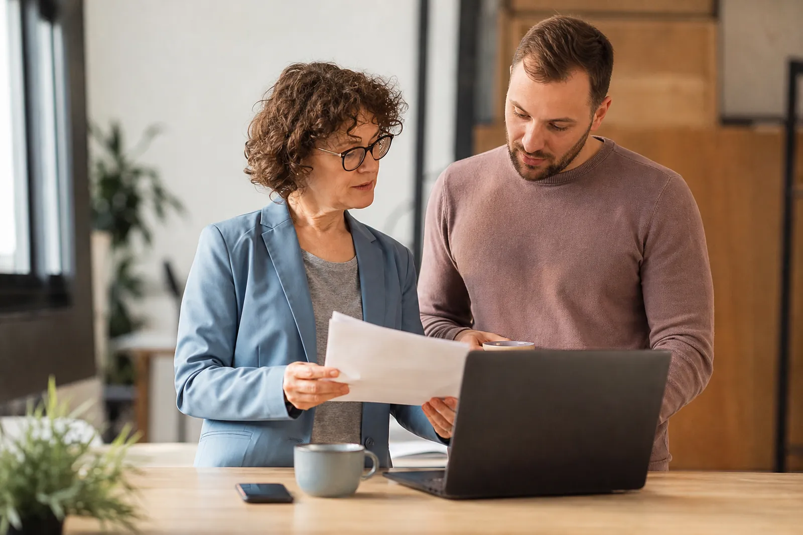 A business owner and a web specialist standing together in a small office, looking at printed notes and a laptop, with a coffee cup and a phone on the desk.