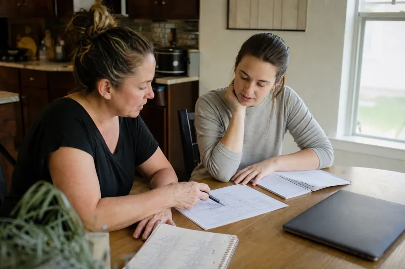 A small business owner and a web helper sit at a kitchen table with printed notes, a laptop closed beside them, and a notepad full of account names, while daylight comes through a 