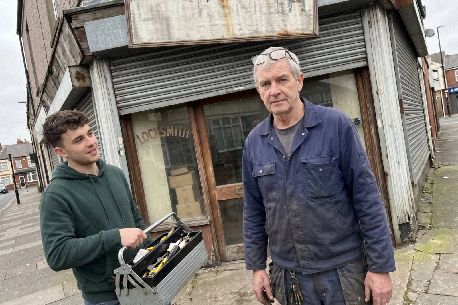 An older locksmith and his apprentice outside their lock and key shop